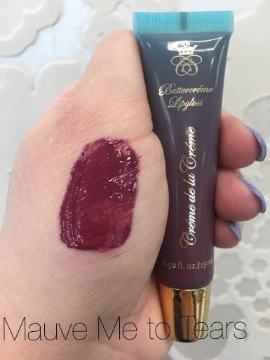 Bold Colours, a hand with a plum-wine lip gloss swatch holding a  lip gloss with gold lid